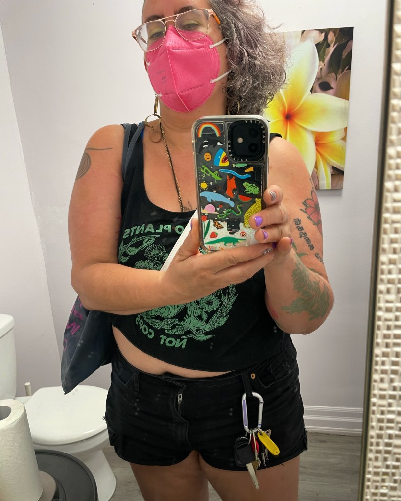 An image of Leah Piepzna-Samarasinha, a light skinned nonbinary femme, doing a mirror selfie: they wear a hot pink KN95 mask, curly silver/brown/lavender hair, and a "talk to plants, not cops" crop top and short black denim shorts.